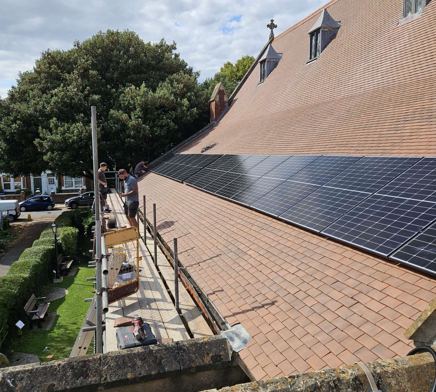 A long run of black solar panels being fitted along a clay-tiled roof, with the installers working from the scaffold beside it